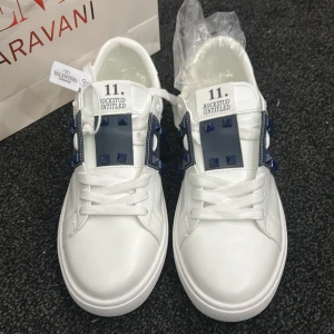 Valentino Rockstud Untitled sneakers vit/blå - Snygga vita Valentino Rockstud Untitled sneakers med blåa detaljer och ikoniska nitar på sidorna. Skorna har snörning, platt sula och är tillverkade i skinn. Perfekt för dig som vill sticka ut med en lyxig och modern look. Priset kan vi prata om