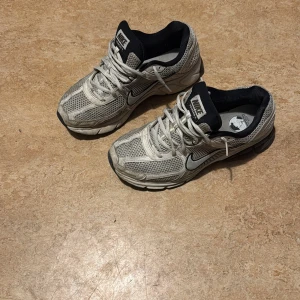Nike vomero 5 - Säljer ett par grå Nike sneakers med silvriga detaljer och svart insida. Skorna har mesh-överdel för bra ventilation, snörning och platt sula. Perfekta för dig som gillar sportig stil och vill ha bekväma skor till vardagen.