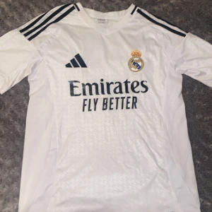 Real Madrid Vini Jr. 7 matchtröja Adidas (falsk) - Säljer en Real Madrid fotbollströja från Adidas i storlek M. Tröjan är vit med svarta detaljer, har korta ärmar och V-ringad krage. Broderat klubbmärke på bröstet, tryck med 'Emirates Fly Better' och Vini Jr. 7 på ryggen. Tillverkad i lätt polyester.