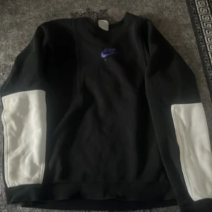 Nike svart sweatshirt med vita detaljer - Svart sweatshirt från Nike med lila logga på bröstet och texten 'GET OVER YOUR FEAR OF HEIGHTS' på ryggen. Tröjan har långa ärmar med breda vita paneler på underarmarna och en klassisk rund hals. Perfekt för en sportig och trendig look.