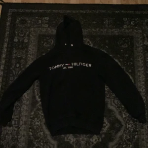 Svart hoodie Tommy Hilfiger - Svart hoodie från Tommy Hilfiger med klassisk logga och texten 'Est. 1985' broderad på bröstet. Tröjan har huva med snörning och långa ärmar. Perfekt för en chill och stilren look.