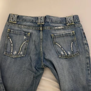 Blå y2k/2000stal bootcut jeans - Snygga medium rise blå jeans. Jeansen har slitna detaljer, nitar på hällor och fickor samt. Materialet är klassisk denim.