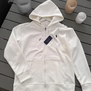 Vit hoodie från Polo Ralph Lauren - Vit hoodie från Polo Ralph Lauren med klassisk svart broderad logga på bröstet. Tröjan har huva med snörning, dragkedja framtill och två stora fickor. Tillverkad i mjuk bomull, perfekt för chill dagar. Stilren och enkel design som funkar året runt.