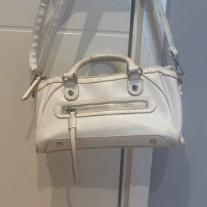 Vit axelväska med dragkedja - Snygg vit axelväska i skinnimitation med både handtag och justerbar axelrem. Väskan har silverfärgade detaljer och en dragkedja framtill. Perfekt storlek för det viktigaste och stilren design som passar till många outfits. Är ganska sliten och därför lågt pris