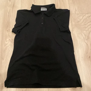 Svart pikétröja från Clique, M - Svart pikétröja från Clique i storlek M. Klassisk design med krage, korta ärmar och tre knappar framtill. Tillverkad i mjuk bomull som känns skön mot huden. Perfekt för dig som gillar enkel och stilren look.