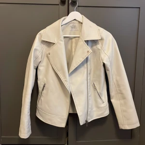 Vit skinnjacka från DeFacto - Snygg vit skinnjacka från DeFacto, bred krage och två fickor med dragkedja. Jackan har en asymmetrisk dragkedja framtill och är långärmad. Mindre märken på ena axeln