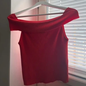 Röd offshoulder topp från Vero Moda - Säljer en snygg röd ribbad topp från Vero Moda i storlek L. Modellen är offshoulder med bred krage som ger en cool och trendig look. Materialet är mjukt och stretchigt, perfekt för att styla med jeans eller kjol. Toppen är ärmlös och har en figurnära passform.