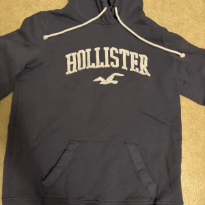 Mörkblå hoodie från Hollister - Snygg mörkblå hoodie från Hollister med vit broderad logga och text på bröstet. Tröjan har huva med vita snören och en klassisk magficka. Perfekt för chill dagar och enkel att matcha med jeans eller joggers.