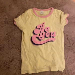 Gul t-shirt med rosa tryck från Lindex - Snygg gul t-shirt från Lindex med coolt rosa och lila tryck 'Be you' på bröstet. T-shirten har rosa kant vid hals och ärmslut, och är gjord i mjuk bomull. Perfekt för dig som gillar färgglada och positiva plagg.