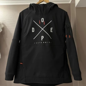 Svart vindjacka från Dope med huva - Svart vindjacka från Dope med stor vit logga framtill, huva med dragsko och röda detaljer på dragkedjorna. Praktisk ficka på ärmen och sportig look. Perfekt för dig som gillar streetwear och vill ha en snygg jacka med coola detaljer. Jackan har 3 fickor och dragkedjor som man kan öppna upp jackan för att vädra/ kyla ner sig under armarna 