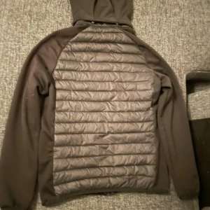 Svart hybridjacka med huva - Snygg svart hybridjacka från Mountain Works med quiltad framsida och rygg, mjuka ärmar och huva. Jackan har dragkedja framtill och två fickor med dragkedja. Perfekt för kyliga dagar och enkel att matcha med allt.