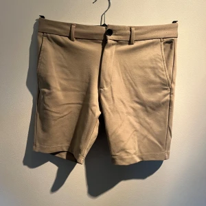 Beige shorts  - Snygga beige shorts med klassisk design, raka ben och bälteshällor. De har två sidofickor fram och två bakfickor med knappdetalj. Perfekta för varma dagar och har en stilren look som funkar till många outfits.