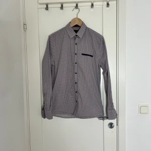 Mönstrad skjorta med blå detaljer - Snygg skjorta med små geometriska mönster i vitt, blått och rött. Skjortan har klassisk krage, lång ärm med manschett och mörkblå knappar. En diskret bröstficka med blå kant ger extra stil. Perfekt för dig som gillar detaljer och färg. Lite slitage i halslinning. Se foto. 