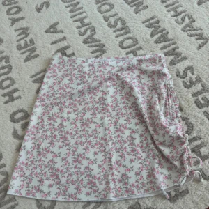 Blommig kortkjol från Gina Tricot - Supersöt vit kortkjol från Gina Tricot med rosa blommönster och snörning på sidan. Kjolen har en lätt A-linjeform och är gjord i mjuk polyester som känns skön mot huden. Perfekt för dig som gillar romantiska och feminina vibes.