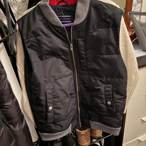 Svart väst från Tommy Hilfiger - Svart quiltad väst från Tommy Hilfiger med grå ribbade muddar vid krage och nederkant. Västen har dragkedja framtill, två fickor med tryckknappar.  Materialet är polyester och insidan har ett stilrent mönster i vitt och marinblått. Bortse från de beiga ärmarna då de är från ett annat plagg.