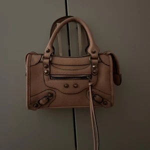 Rosa handväska med dragkedja - Snygg rosa handväska i skinn med coola metalldetaljer och dekorativa nitar framtill. Väskan har två stadiga handtag och en praktisk dragkedja upptill samt en extra ficka med dragkedja på framsidan. Perfekt storlek för det viktigaste och stilren design.