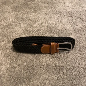 Svart flätat bälte med skinn-detaljer - Snyggt svart flätat bälte med bruna skinn-detaljer och silvrigt spänne. Perfekt för att ge din outfit en stilren touch. Bältet har en klassisk form och passar till både jeans och chinos. Flätad design gör det enkelt att justera längden.