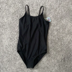 Svart baddräkt med smala axelband - Helt ny inte ens andvänd svart baddräkt med smala axelband och enkel design. Perfekt för dig som gillar klassisk look på stranden eller vid poolen. Materialet är mjukt och följsamt, vilket gör den bekväm att bära hela dagen. Passar dig som vill ha en tidlös och snygg baddräkt.