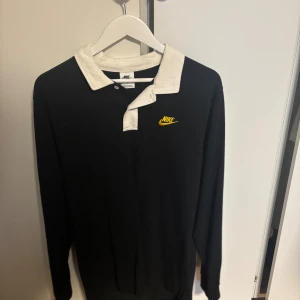 Svart pikétröja från Nike med tryck - Svart långärmad pikétröja från Nike med vit krage och gul Nike-logga på bröstet. På ryggen finns ett stort tryck med siffran 72 i grått och en gul Nike-logga ovanpå. Perfekt för dig som gillar sportig och clean stil.