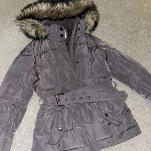 ESPRIT pufferjacka med päls - Grå pufferjacka från ESPRIT med snyggt bälte i midjan och fuskpäls på huvan. Jackan har quiltad insida, långa ärmar med muddar, flera fickor med knappar och dragkedja. Perfekt för kalla dagar.