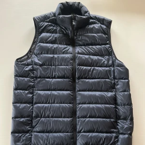 Blå dunväst från Uniqlo, S - Snygg svart dunväst från Uniqlo i storlek S. Västen har quiltad design med horisontella sömmar, hög krage och dragkedja framtill. Tillverkad i lätt och vattenavvisande polyester med dunfyllning. Perfekt för lager-på-lager under kyliga dagar.