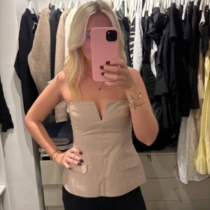 Beige korsett-topp med v-ringning - Säljer en beige korsett-topp med snygg v-ringning framtill och breda axelbandslösa kanter. Toppen har diskreta fickdetaljer och ett lätt ribbat mönster. Helt ny