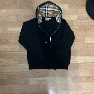 Svart hoodiejacka från Burberry - Svart hoodiejacka från Burberry med klassisk rutig huva i beige, rött och svart. Jackan har dragkedja framtill och snörning vid huvan. Perfekt för dig som vill ha en stilren look med lyxig touch och ikoniskt Burberry-mönster.