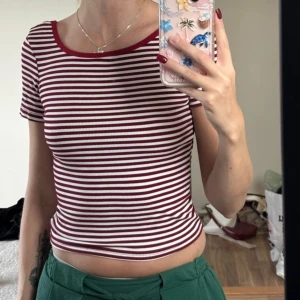 Randig croppad topp med öppen rygg - Säljer en croppad topp med röda och vita ränder, ribbad struktur och korta ärmar. Toppen har en snygg öppen rygg och rundad halsringning med röd kant. Perfekt för dig som gillar en enkel men cool stil.
