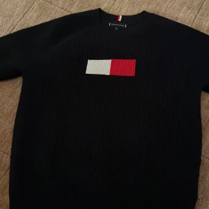 Svart stickad tröja Tommy Hilfiger - Svart stickad tröja från Tommy Hilfiger med klassiskt rött och vitt broderat flaggmärke på bröstet. Rund hals och ribbade muddar. Snygg detalj med röd och vit rand vid nederkanten. Perfekt för dig som gillar stilrena och ikoniska plagg.