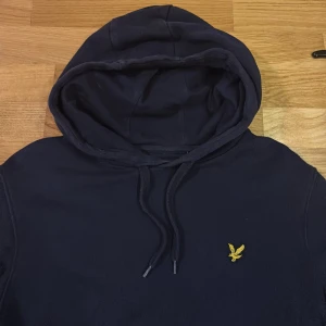 Marinblå hoodie från Lyle & Scott - Snygg marinblå hoodie från Lyle & Scott med klassisk gul broderad fågel på bröstet. Tröjan har huva med snörning, magficka och långa ärmar. Perfekt för chill dagar och enkel att matcha med jeans eller joggers.