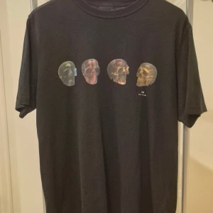 Svart t-shirt med döskalletryck Paul Smith L - Svart t-shirt från Paul Smith med coolt tryck av fyra döskallar i olika färger på bröstet. Klassisk rund hals och korta ärmar. Materialet är mjuk bomull som känns skön mot huden. Perfekt för dig som gillar unika prints och streetstyle.