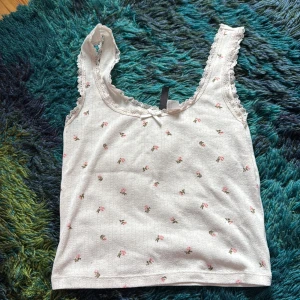 Vit blommig linnetopp med spets - Supersöt vit linnetopp med små rosa blommor och spetskant runt axelbanden. Toppen har en liten rosett framtill och är ärmlös med en mjuk, feminin look. Perfekt för dig som gillar romantisk stil och vill ha något lätt och somrigt.