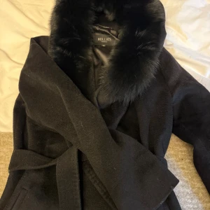 Svart kappa med päls från Hollies - Snygg svart kappa från Hollies med bred krage i pälsimitation och knytskärp i midjan. Kappan har lång ärm och är tillverkad i mjukt material med slät insida. Perfekt för dig som vill ha en stilren och lyxig look under kalla dagar.