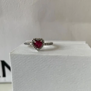 Pandora silverring med röd hjärtsten - Snygg ring från Pandora i äkta silver med en röd hjärtformad sten i mitten, omgiven av små glittrande stenar. Ringen har ett detaljerat band med pärlstruktur som ger extra bling. Perfekt för dig som gillar romantiska och trendiga smycken.
