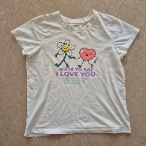 T-shirt - Vit t-shirt från Wow i mjuk bomull med korta ärmar. Framsidan har ett färgglatt tryck med en glad blomma och ett hjärta som håller hand, samt texten 'Ways to say I love you' i lila. Rund hals och avslappnad passform. Perfekt för dig som gillar gulliga motiv. Finns en fläck.