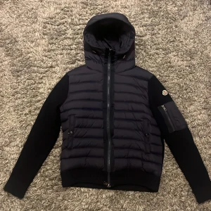 Svart Moncler pufferjacka med huva - Svart pufferjacka från Moncler med stickade ärmar och huva. Jackan har dragkedja framtill, ribbade muddar och Moncler-logga på ärmen. Fickor med tryckknappar och en extra ficka med dragkedja på ärmen. Perfekt för kalla dagar och riktigt snygg streetstil.