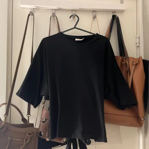 Svart topp med knyt från Nelly - Svart topp från Nelly med rund halsringning och vida, korta ärmar. Toppen har en snygg knytning i ryggen som ger en cool detalj. Tillverkad i mjuk bomull, perfekt för en avslappnad och trendig look. 💕