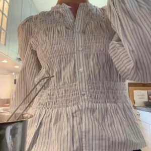 Randig blus med smock och puffärm - Säljer en vit och grårandig blus med smockdetaljer över bröst och midja. Blusen har knappar framtill, v-ringning och långa puffärmar. Perfekt för dig som gillar en luftig och trendig look. Jag har sytt ditt en extra innerknapp som inte syns utåt men som gör att inte bhn syns i ett glapp mellan knapparna