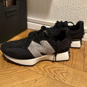Svarta New Balance sneakers med leoparddetalj - Snygga svarta New Balance sneakers med vit sula och stor N-logga i svartvitt leopardmönster på sidan. Skorna har snörning, rund tå och är tillverkade i en mix av textil och mocka. Perfekta för dig som vill sticka ut med en cool detalj på fötterna. Endast använda ett fåtal gånger och skicket är 10/10!