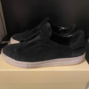 Svarta sneakers från Axel Arigato - Svarta sneakers från Axel Arigato med stilren design och vit platt sula. Skorna är handgjorda i Portugal och har en ovandel i mocka med diskret logga på sidan. Insidan är fodrad med mjukt skinn för extra komfort. Perfekta för dig som gillar minimalistisk stil.