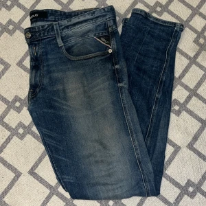 Blå Replay jeans, modell 34/32 - Säljer ett par snygga blå jeans från Replay i klassisk femficksmodell. Jeansen har en lätt tvättad look med diskreta slitningar och raka ben. Märkesdetaljer på ficka och bak, samt tydlig Replay-logga i midjan. Perfekta för dig som gillar stilrena jeans med lite edge. 