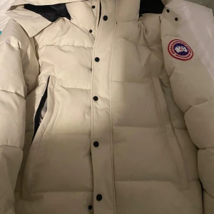 Beige pufferjacka från Canada Goose - Säljer en beige pufferjacka från Canada Goose med ikonisk patch på ärmen. Jackan har huva, svarta knappar och dragkedja, samt praktiska fickor. Perfekt för kalla dagar med sin quiltade design och tjocka dunfyllning. Stilsäker och varm jacka för vintern.