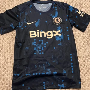 Chelsea FC mörkblå matchtröja Nike - Chelsea FC fotbollströja i mörkblått med ljusblå digitalt inspirerat mönster. Tröjan har korta ärmar, rund hals och BingX sponsortryck framtill. Tillverkad i lätt och ventilerande material, perfekt för träning eller match.