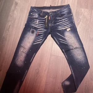 DSQUARED2 blå jeans med dragkedja - Snygga blå jeans från DSQUARED2 med coola slitningar, broderade detaljer och färgstänk. Framtill finns en unik dragkedja och patchar som ger ett edgy uttryck. Klassisk femficksmodell med DSQ2-logga bak. Passformen är smal och materialet är jeans.
