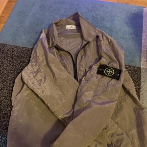 Grå overshirt från Stone Island - Säljer en grå overshirt från Stone Island med klassisk patch på ärmen. Jackan har två stora bröstfickor med lock och en svart dragkedja framtill. Materialet är lätt och syntetiskt, perfekt för lager-på-lager. Stilren och modern look.