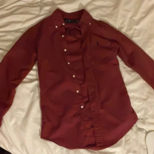 Vinröd skjorta från Ralph Lauren XS - Vinröd skjorta från Ralph Lauren i slim fit med klassisk krage och vita knappar. Broderad logga på 