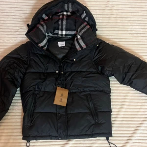 Svart pufferjacka från Burberry M - Svart pufferjacka från Burberry med avtagbara ärmar, huva och klassiskt rutigt foder. Jackan har dragkedja, knappar och två fickor med dragkedja. Materialet är bomull med detaljer i viskos och polyamid. Perfekt för kalla dagar med snygg streetkänsla.