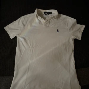 Vit polo från Ralph Lauren slim fit - Vit klassisk polo från Ralph Lauren i slim fit-modell. Skjortan har korta ärmar, krage och knappar vid halsen. Ikoniska broderade loggan på bröstet. Tillverkad i mjuk bomull som är skön mot huden och passar perfekt till jeans eller shorts.