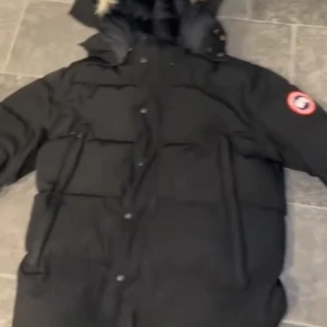 Svart dunjacka från Canada Goose L - Säljer en svart dunjacka från Canada Goose med klassisk patch på ärmen och stor huva med fuskpäls. Jackan har flera fickor, dragkedja och knappar framtill. Perfekt för kalla vinterdagar och riktigt snygg streetstil.priset kan diskuteras!!!