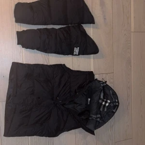 Svart dunväst från Burberry med huva - Svart dunväst från Burberry med avtagbara ärmar och huva. Huva med klassiskt rutigt Burberry-mönster i grått, vitt och svart. Västen har dragkedja och tryckknappar framtill samt fickor med dragkedja. Märke 'Burberry London England' på ena ärmen.